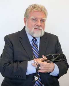 A photograph of Dr. John Trowbridge smiling in a dark suit holding a stethoscope. 